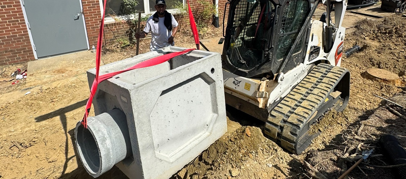 Skid-steer lowers precast concrete drainage structure at construction site