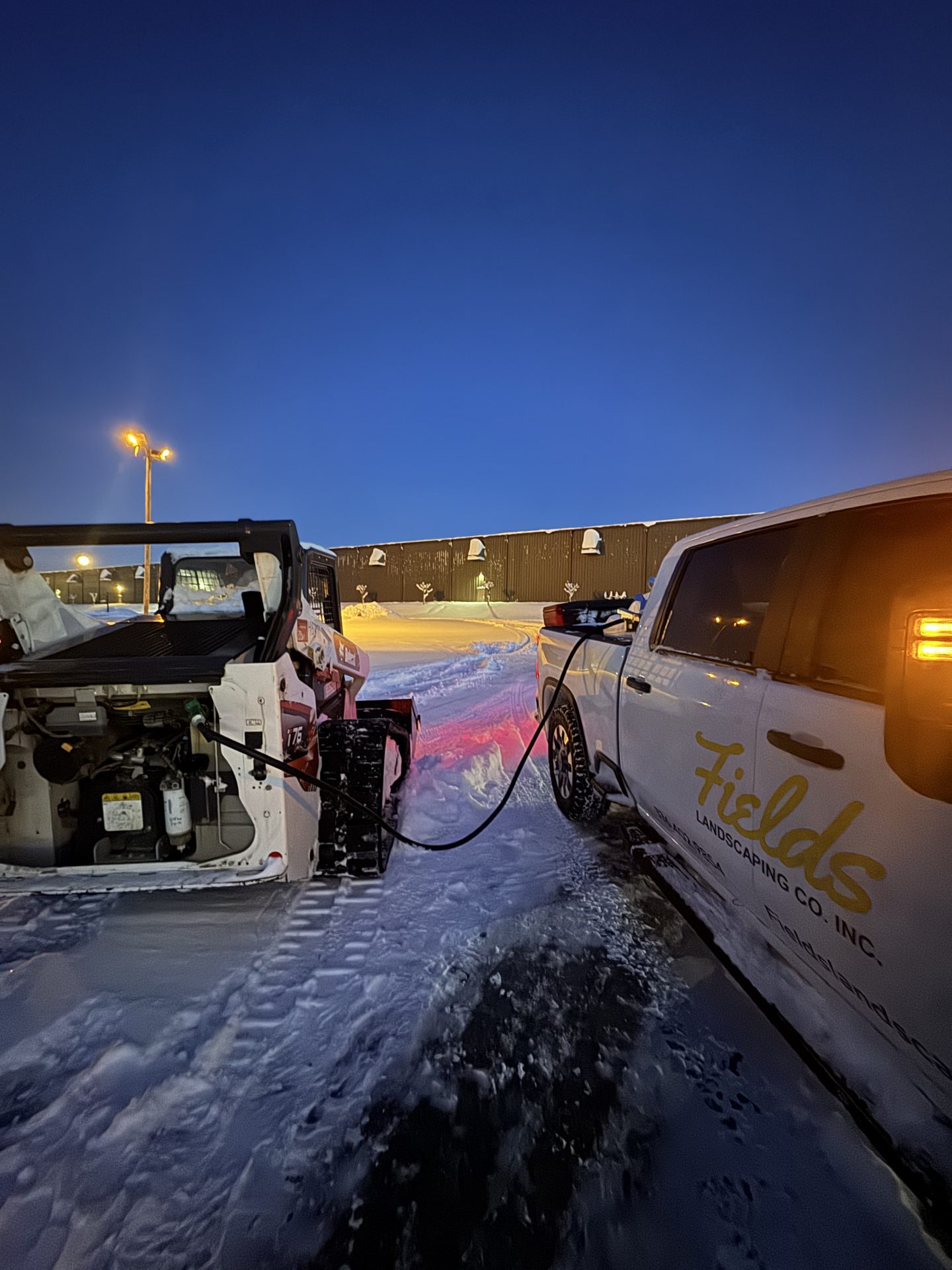 Landscaping truck jump-starting snow-covered skid steer at dawn